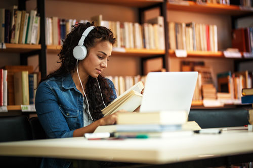 A woman sits in a library, working on her laptop with headphones on.