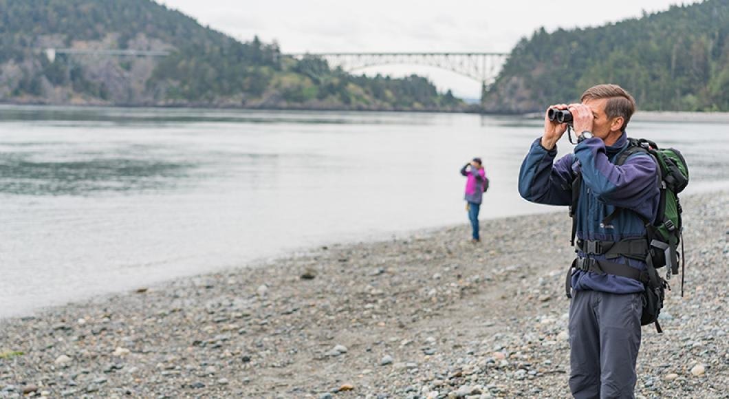 Man using binoculars on the beach. In the background the Deception Pass bridge spans Fidaglo island from the mainlain.