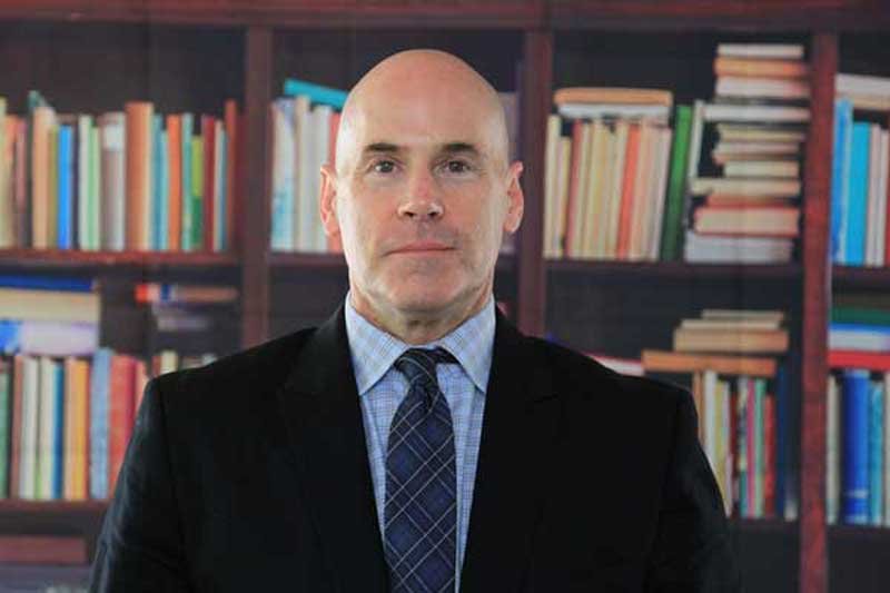 Anthony, a clean shaven man in a suit and tie, stands in front of a fully loaded shelf of books.