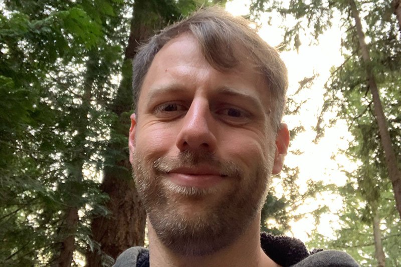 Andrew sporting a trimmed beard and mustache takes a selfie in the deeply forested woods.