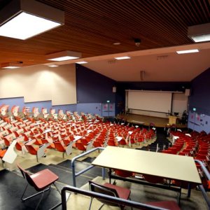 Emply lecture hall filled with rows of seats facing a stage with a screen behind it.