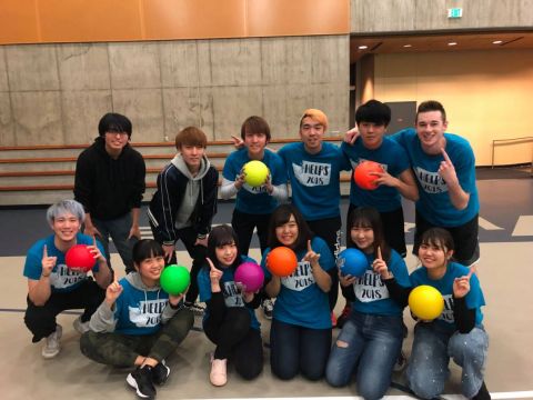 Group of WWU-AUAP students hold brightly colored balls, wearing matching blue sport t-shirts in a gymnasium, smiling for the camera