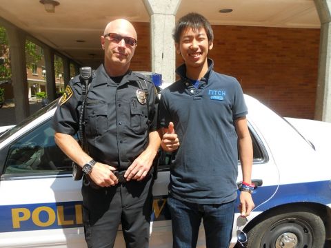 An AUAP student stands next to a police officer on campus, leaning against a police car.