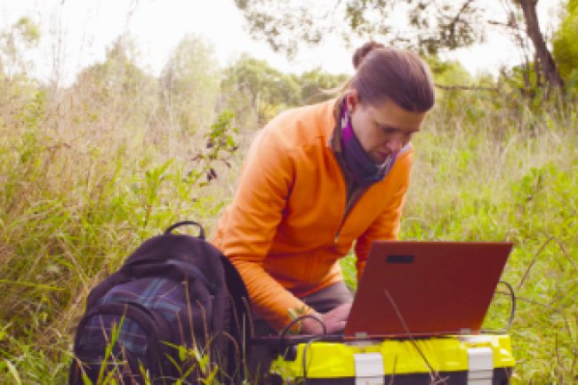 A person working in the field of  Data Science crouches outdoors, typing in data on a laptop computer