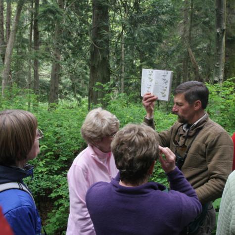 a group of students in the lush green forest stand listening to a speaker, who is holding up an identification guide